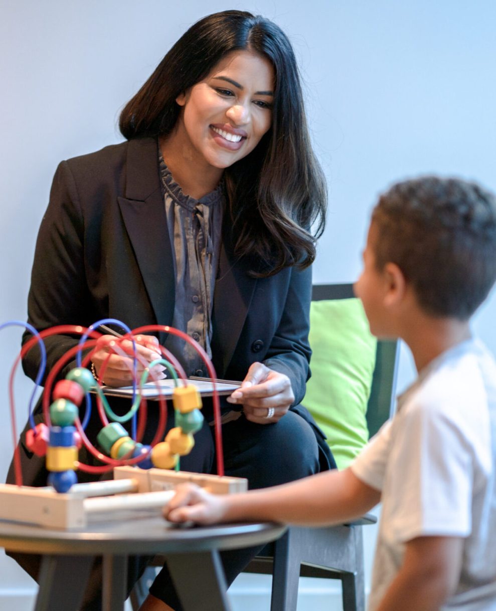 A female therapist sits in a chair beside her young patient as they play together with a bead maze. The therapist is dressed professionally in a suit and has a tablet out on her lap to take notes about the session. The young causally dressed boy is kneeling on the floor n front of the toy as they talk together.